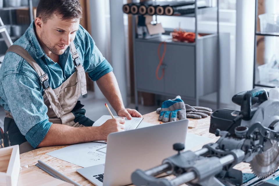 Atelier menuiserie intérieur avec artisan en salopette travaillant sur ordinateur portable, prenant notes avec crayon sur plans techniques
