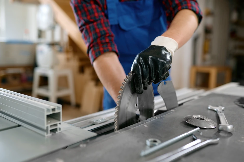 Atelier de menuiserie intérieur avec artisan en bleu de travail et gants de protection noirs manipulant une scie circulaire sur table