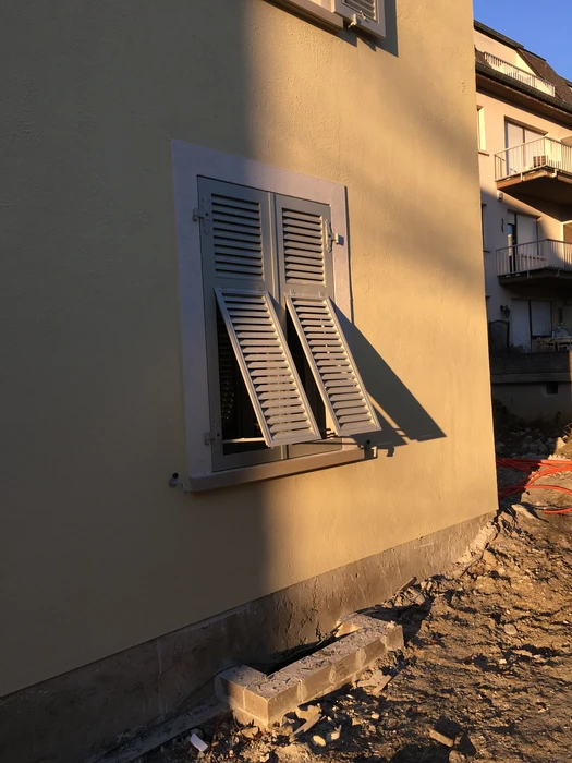 Façade extérieure d'un bâtiment en cours de construction, mur en enduit jaune ocre, fenêtre équipée de volets battants à lames persiennées gris clair, ouverts à 90°