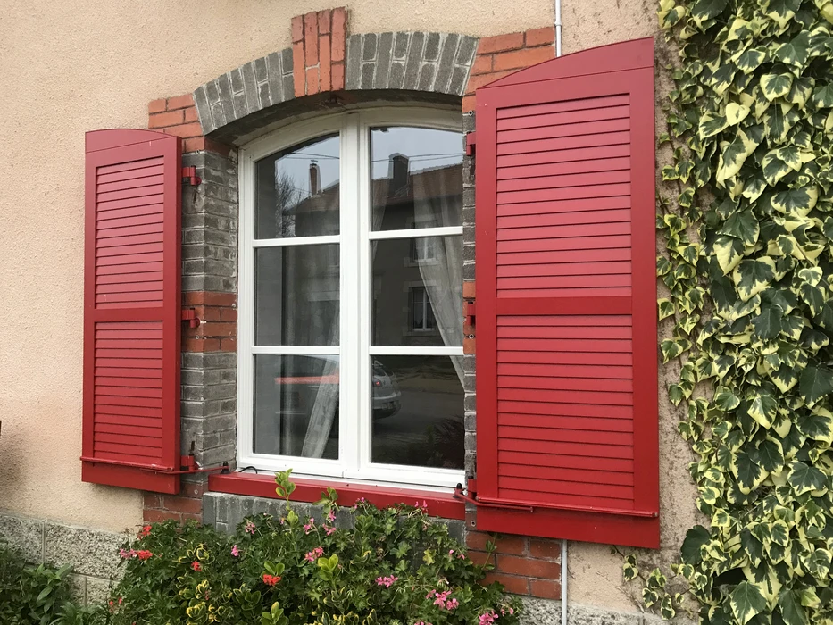 Façade de maison avec fenêtre à arc cintré, volets battants à lames orientables peints en rouge vif