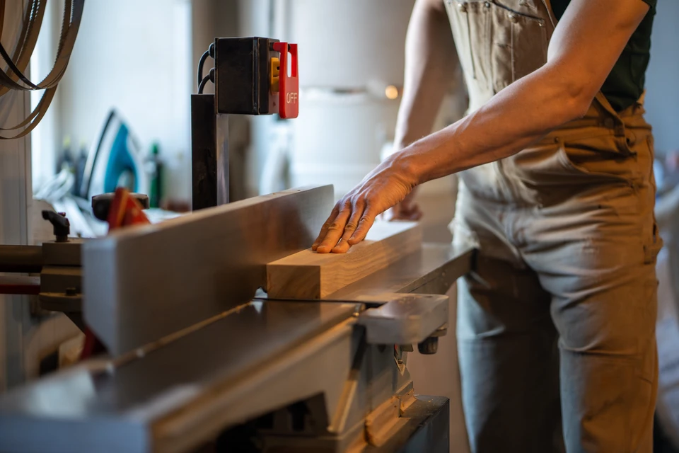 Femme travaillant dans un atelier de menuiserie lumineux, portant salopette en jean et chemise à carreaux, utilisant un marteau sur une pièce de bois