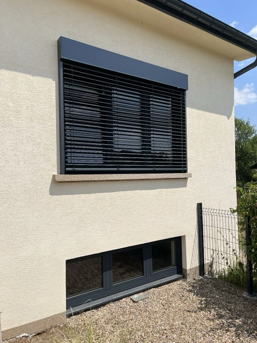Terrasse couverte extérieure attenante à une maison individuelle, équipée de volets roulants aluminium couleur blanc cassé, encadrés de profilés anthracite