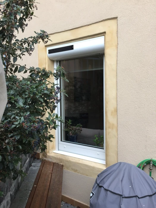 Façade extérieure d'un bâtiment en cours de construction, mur en enduit jaune ocre, fenêtre équipée de volets battants à lames persiennées gris clair, ouverts à 90°