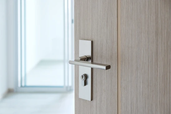 Intérieur résidentiel moderne avec porte coulissante en bois clair partiellement ouverte, poignée encastrée design ovale, rideaux beiges avec embrasses, vue sur extérieur verdoyant